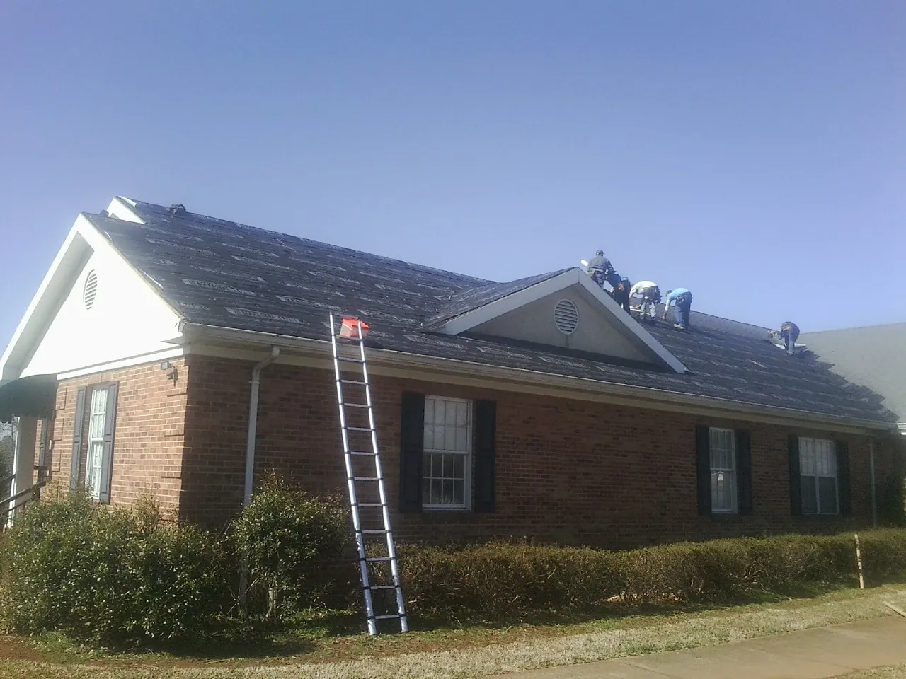 Crew tearing shingles off a brick ranch roof with a ladder set — Metal Roof Repair work by our crew in Oronoco