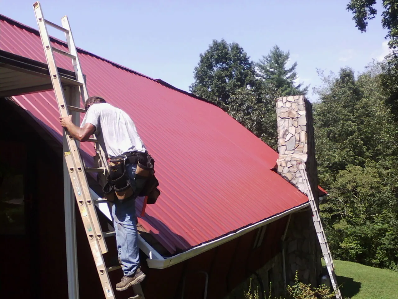 Roofer on a ladder beside a red metal roof and stone chimney — Soffit & Fascia Repair by Roofing Brothers in Oronoco
